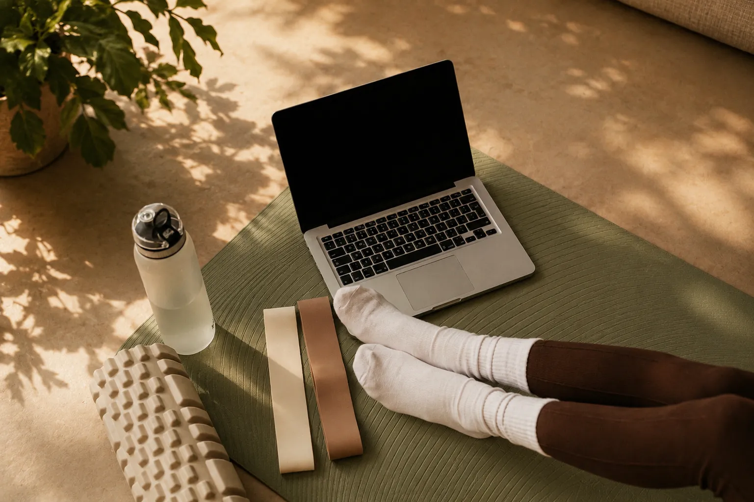 Person auf einer Yogamatte mit Laptop, Widerstandsbändern, Trinkflasche und Schaumstoffrolle