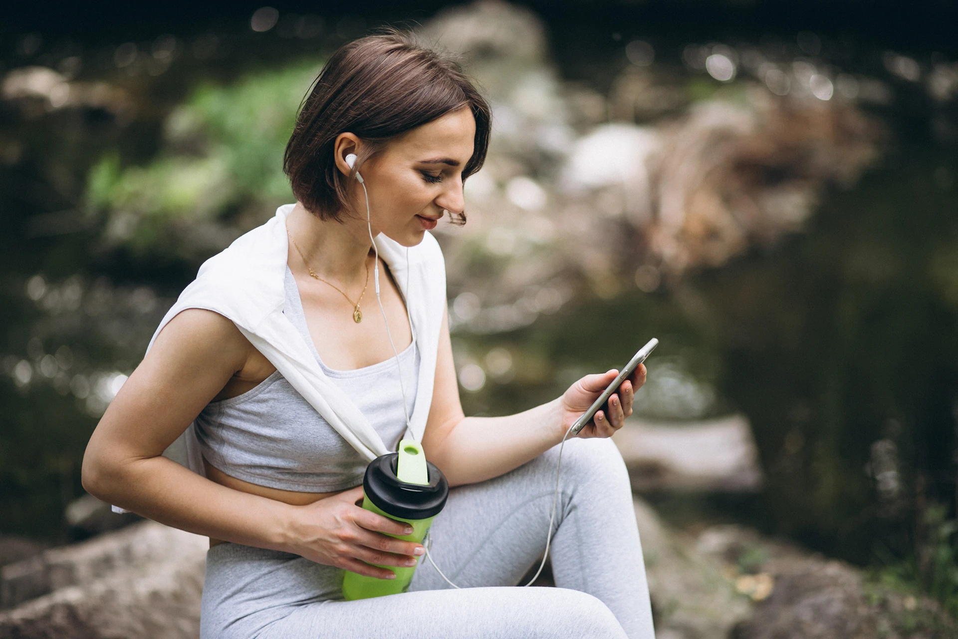 Junge Frau in Sportkleidung sitzt im Freien, hört mit Ohrhörern Musik und schaut auf ihr Smartphone, mit einer Trinkflasche in der Hand